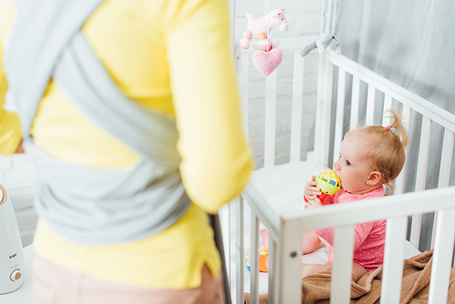 Selective focus of infant girl biting toy in crib near mother with sling at home