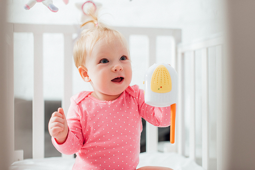 Selective focus of excited infant girl with baby monitor looking away in crib