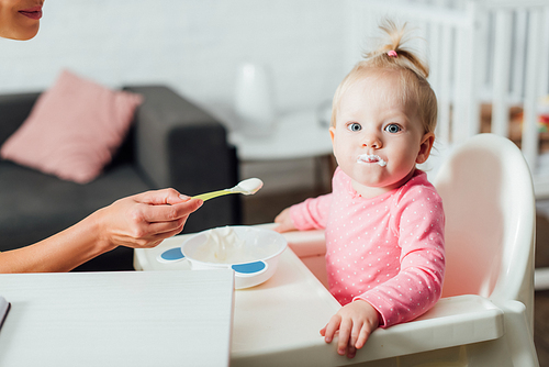 Selective focus of infant girl with messy mouth  near mother with spoon of baby food