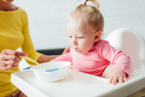 Selective focus of infant girl sitting in highchair near mother with spoon and bowl of baby food