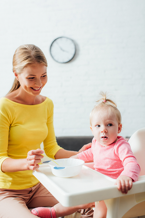 Selective focus of mother looking at baby girl near bowl and spoon on feeding chair