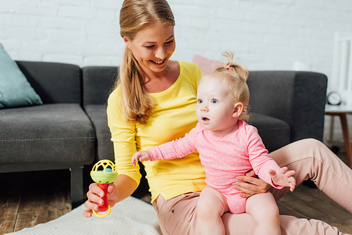 Mother holding rattle and excited baby daughter on floor at home