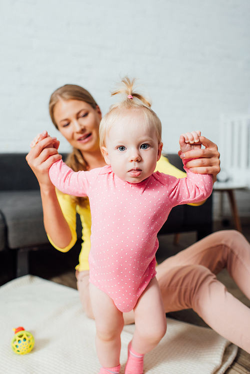 Selective focus of woman holding hands of infant daughter walking at home