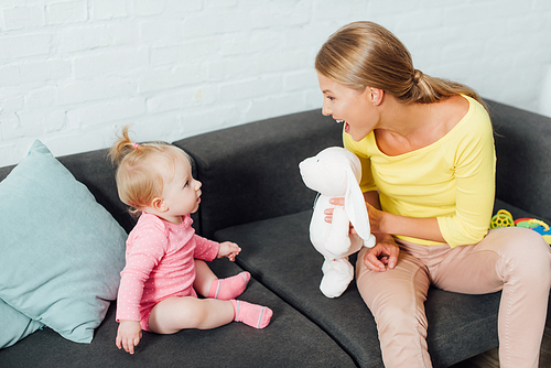 Mother holding soft toy near infant daughter on sofa