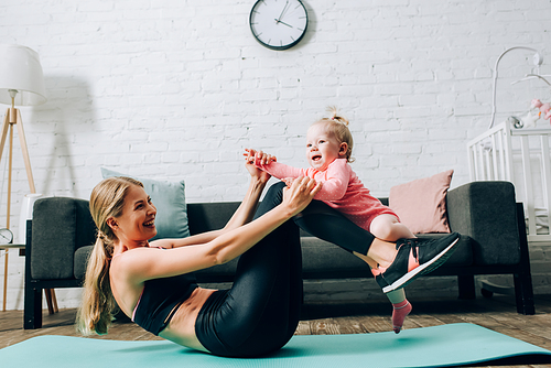 Woman in sportswear holding infant daughter while exercising on fitness mat in living room