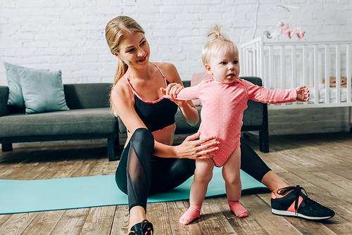 Sportswoman holding infant daughter while sitting on fitness mat in living room