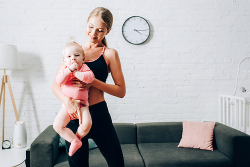 Fit woman in sportswear holding baby daughter on hands in living room