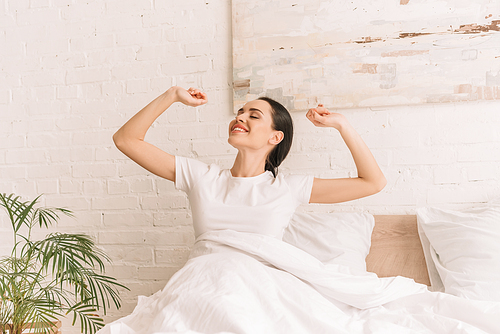 pretty, smiling woman stretching with closed eyes while sitting in bed under blanket