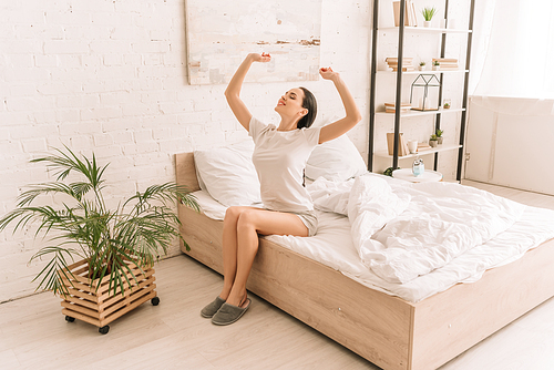 smiling woman in pajamas stretching with closed eyes while sitting on bed near potted plant