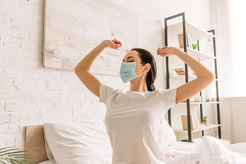 young woman in pajamas and medical mask stretching hands in bedroom