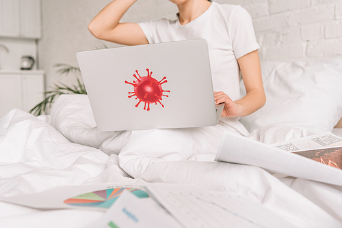 cropped view of freelancer working in bed on laptop with coronavirus bacteria sticker