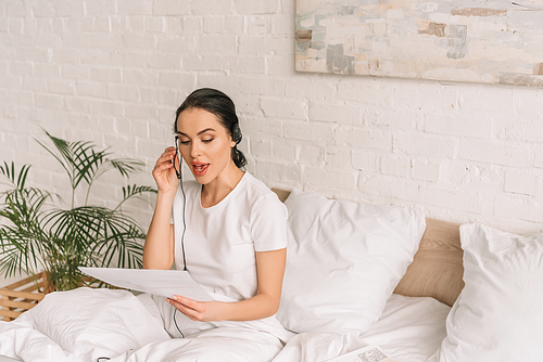 attractive remote operator in headset holding paper while working in bed