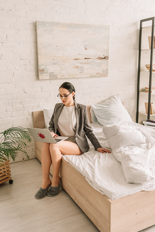 attractive businesswoman in blazer over pajamas using laptop while sitting on bed