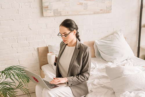 attentive businesswoman in blazer over pajamas using laptop and holding cup of coffee in bedroom
