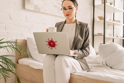 attractive businesswoman in blazer over pajamas using laptop and holding cup of coffee while sitting on bed