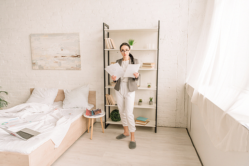 young businesswoman in blazer over pajamas holding papers while  in bedroom