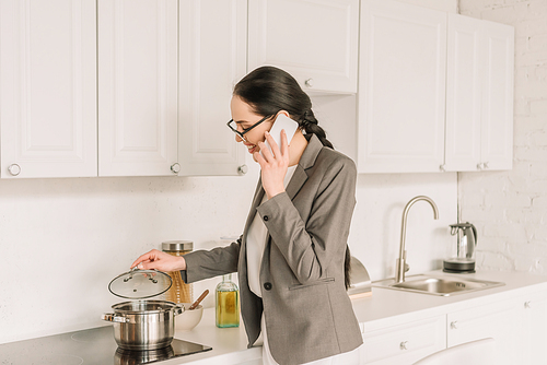 smiling businesswoman in blazer over pajamas cooking and talking on smartphone in kitchen