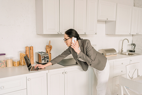 businesswoman in blazer over pajamas talking on smartphone and using laptop in kitchen