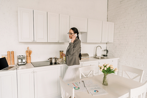 businesswoman in blazer over pajamas talking on smartphone while cooking in kitchen near table with tulips and documents