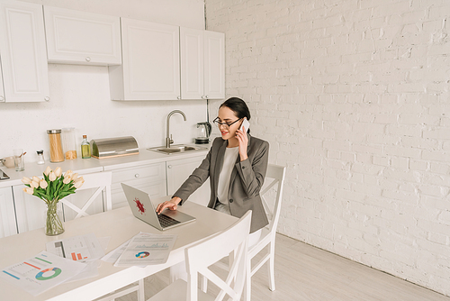 smiling businesswoman in blazer over pajamas talking on smartphone and using laptop in kitchen near documents and fresh tulips
