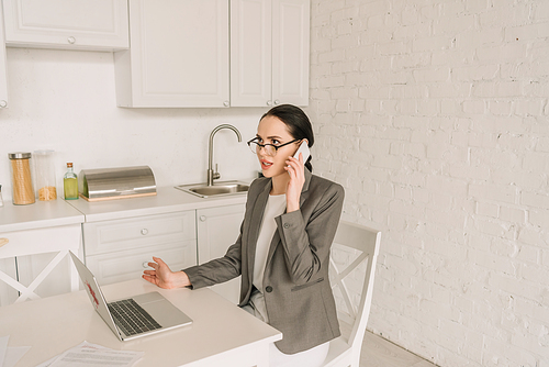 young businesswoman in blazer over pajamas talking on smartphone while working in kitchen near laptop