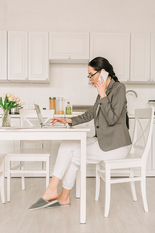 side view of businesswoman in blazer over pajamas working in kitchen, talking on smartphone and using laptop
