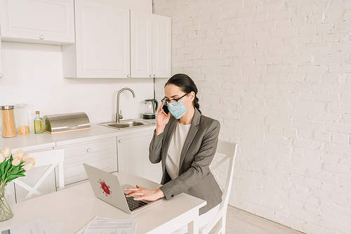 businesswoman in medical mask and blazer over pajamas working in kitchen, talking on smartphone and typing on laptop