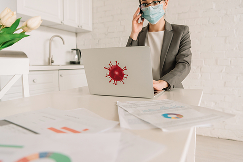 selective focus of shocked businesswoman in medical mask talking on smartphone near laptop with coronavirus bacteria sticker