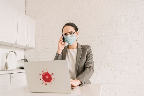 serious businesswoman in medical mask and eyeglasses talking on smartphone near laptop with coronavirus bacteria sticker