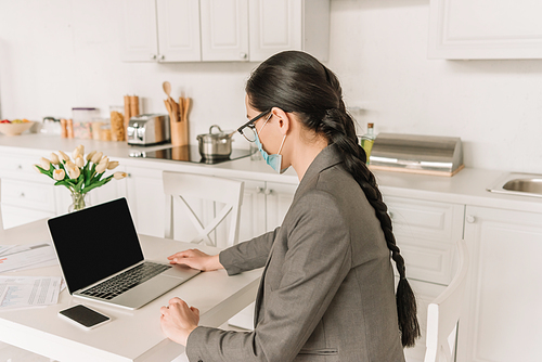 young brunette businesswoman in medical mask working on laptop in kitchen