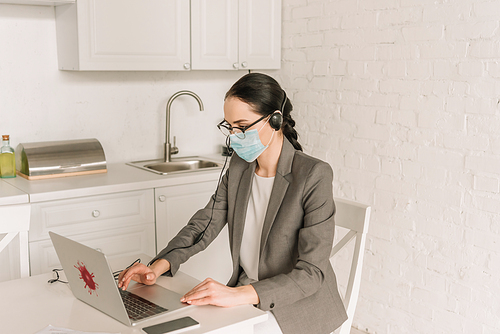 young businesswoman in medical mask typing on laptop with coronavirus sticker in kitchen