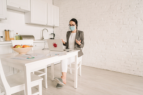 businesswoman in medical mask and blazer over pajamas gesturing while working at kitchen near laptop