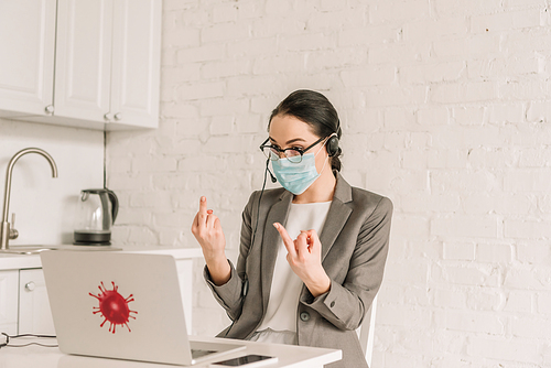 angry businesswoman in medical mask and headset showing middle fingers while working in kitchen near laptop