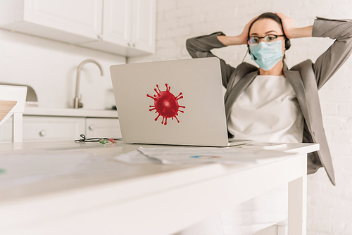 selective focus of tired businesswoman in medical mask and headset touching head while looking at laptop with bacteria sticker in kitchen