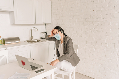 tired businesswoman in blazer over pajamas, medical mask and headset sitting near laptop while working at home