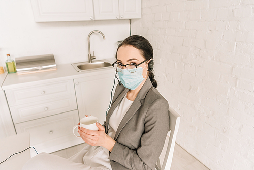 young businesswoman in medical mask, headset and blazer holding cup of coffee in kitchen