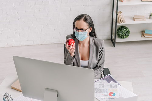 high angle view of businesswoman in medical mask holding ripe apple while working at home near computer monitor