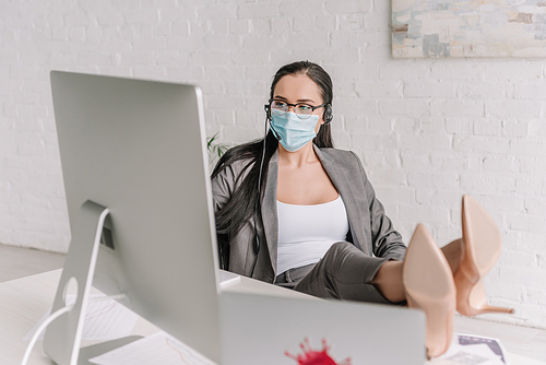 selective focus of businesswoman in medical mask and headset working at home while holding legs on table