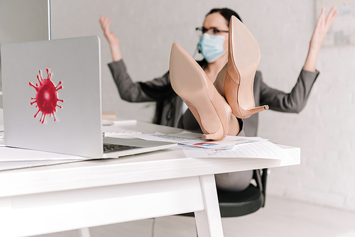 selective focus of businesswoman in medical mask showing shrug gesture while working at home with legs on table
