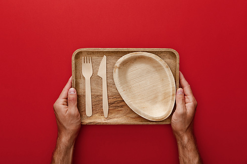 cropped view of man holding natural rectangular wooden dish with plate, fork and knife on red background