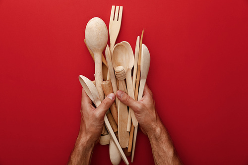 cropped view of man holding wooden spoons, fork and kitchenware on red background