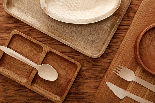 top view of wooden dish and cutting board with plates and cutlery on brown background