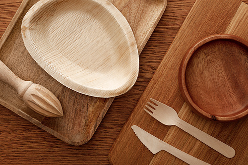top view of wooden dish and cutting board with plates, cutlery and hand juicer on brown background