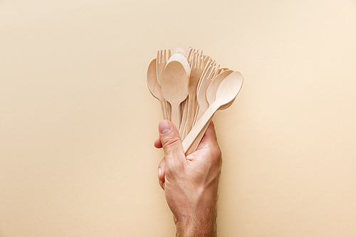 cropped view of man holding natural wooden spoons and forks on beige background