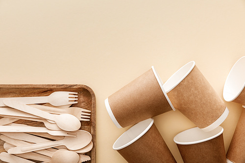 top view of rectangular wooden dish with forks and spoons near paper cups on beige background