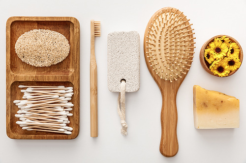 flat lay with wooden dish with cotton swabs and loofah near toothbrush, hairbrush, piece of soap, pumice stone and cup with flowers on white background