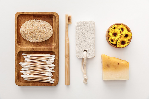 flat lay with wooden dish with cotton swabs and loofah near toothbrush, piece of soap, pumice stone and cup of flowers on white background