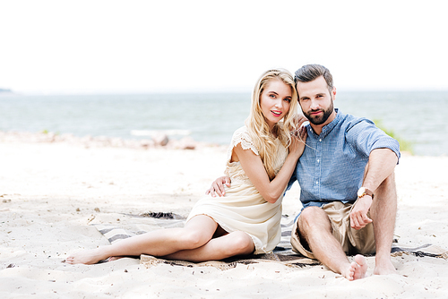 beautiful young barefoot couple sitting on blanket at beach near sea