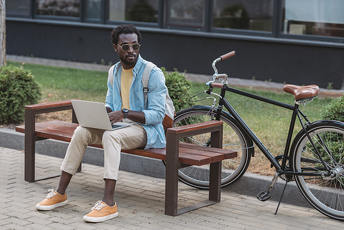 handsome african american man using laptop while sitting on bench near bicycle and looking away