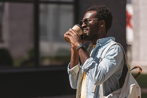 cheerful african american man in sunglasses drinking coffee from paper cup while talking on smartphone